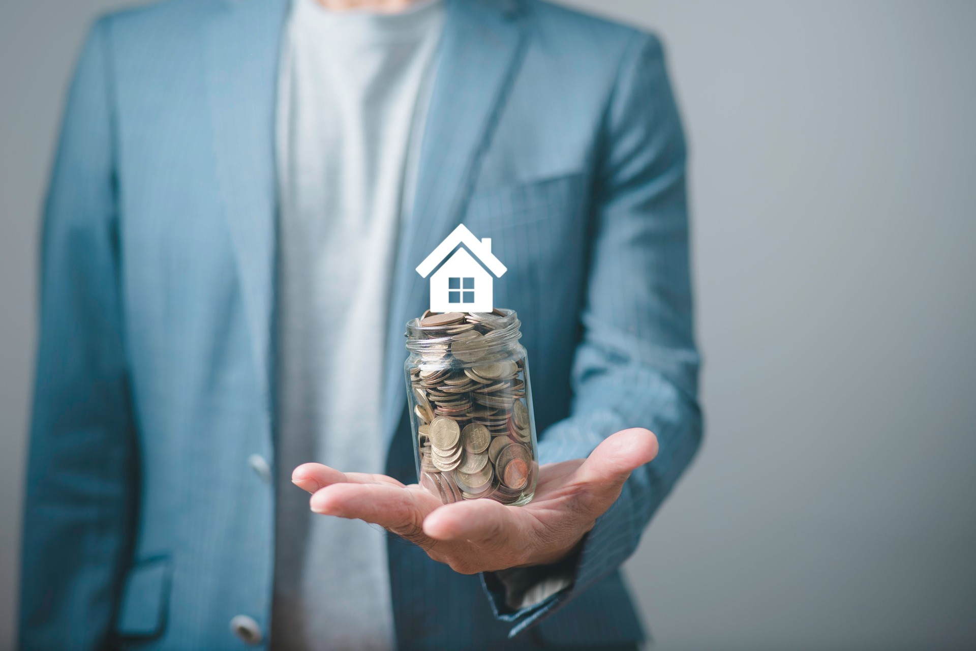 Businessman holding a jar full of coins with a house icon above it, symbolizing real estate investment, home savings, financial planning, mortgage, and property investment concept.